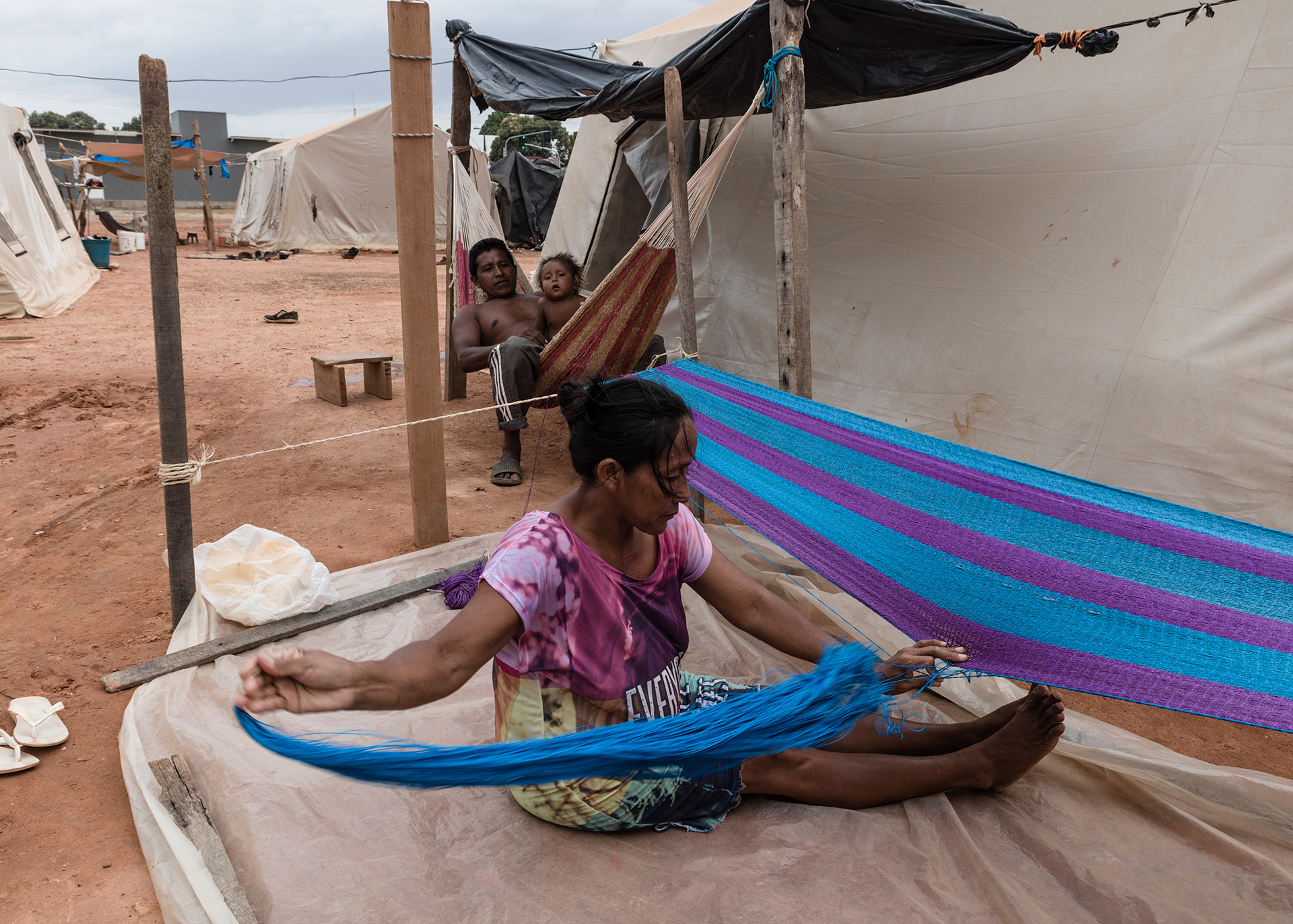 At Pintolandia shelter, an indigenous woman sewing a hammock while her wife take care of their baby, 2019.